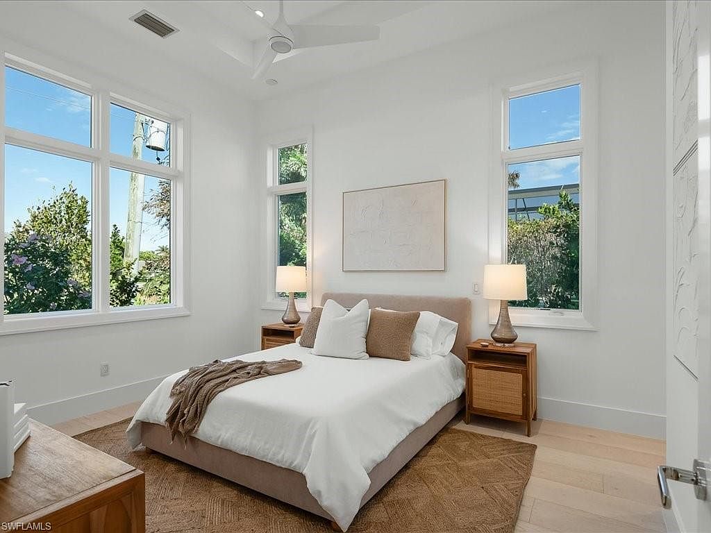 This is a bright and airy primary bedroom featuring a neutral color palette. The room is well-lit with natural light streaming through multiple windows, highlighting the hardwood floors and a comfortable-looking bed with white linens and decorative pillows. Two matching nightstands with lamps flank the bed, adding symmetry and functionality to the space.