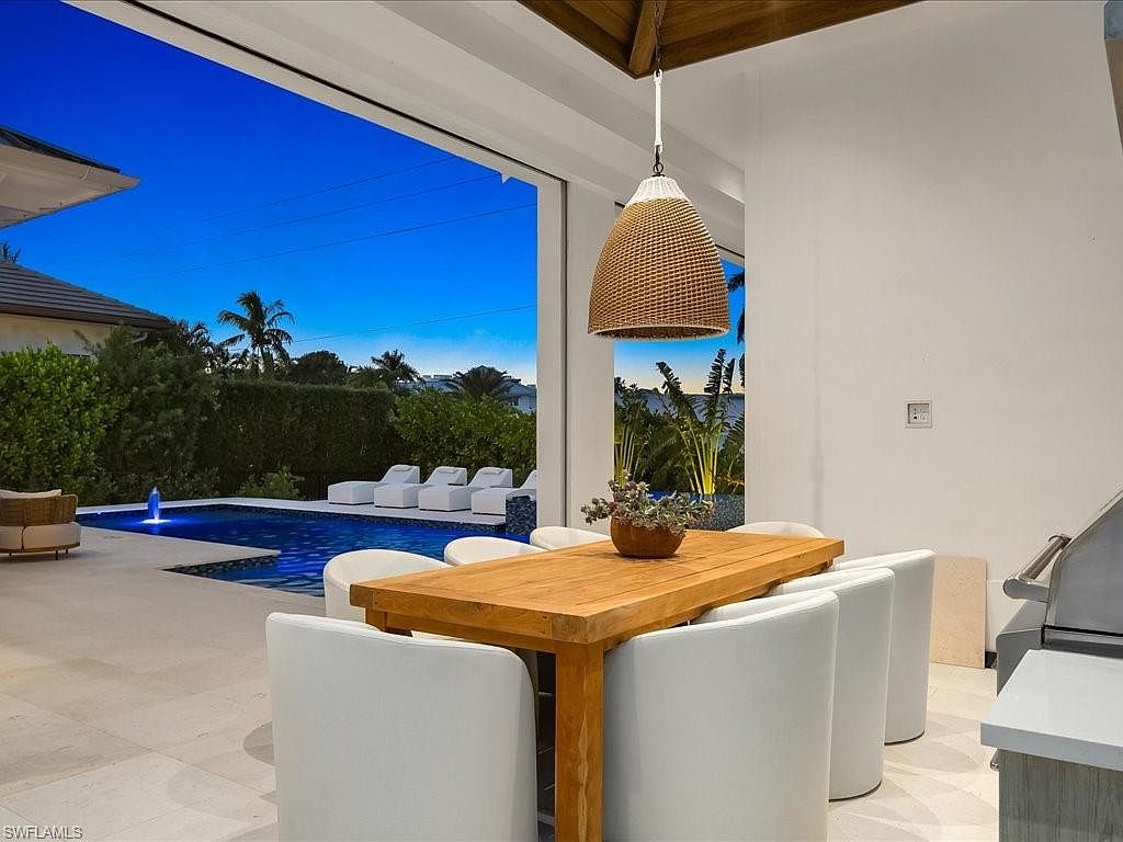 This image showcases an elegant outdoor dining area adjacent to a luxurious pool. The space features a wooden dining table surrounded by white upholstered chairs, illuminated by a stylish pendant light. The pool area includes lounge chairs and lush landscaping, creating a serene and inviting atmosphere perfect for relaxation and entertaining.