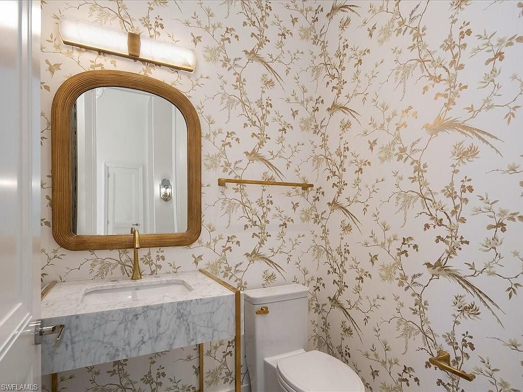 This is a stylish guest bathroom featuring a marble countertop vanity with a gold faucet and a matching gold-framed mirror. The walls are adorned with a delicate bird and floral wallpaper, complemented by gold fixtures like the towel rack and toilet paper holder. The overall impression is elegant and sophisticated, creating a luxurious powder room experience.
