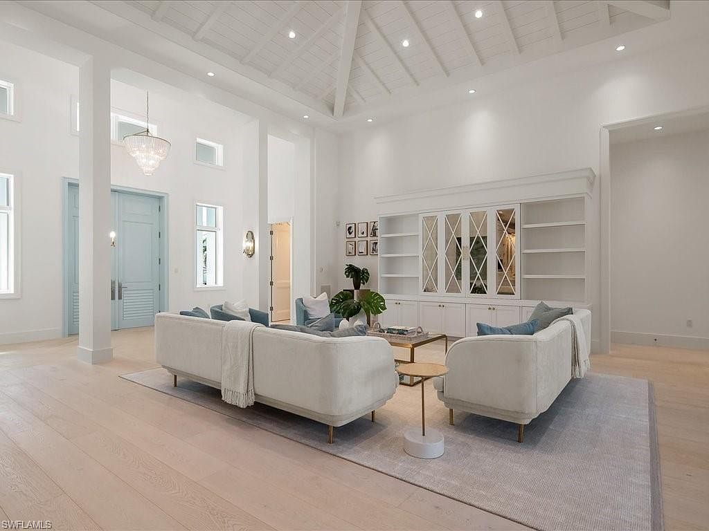 This is a bright and spacious living room featuring high ceilings with exposed beams, white walls, and light wood flooring. The room is furnished with two cream-colored sofas, a light gray rug, and a built-in shelving unit with mirrored doors. A chandelier hangs elegantly, adding a touch of sophistication to the space.