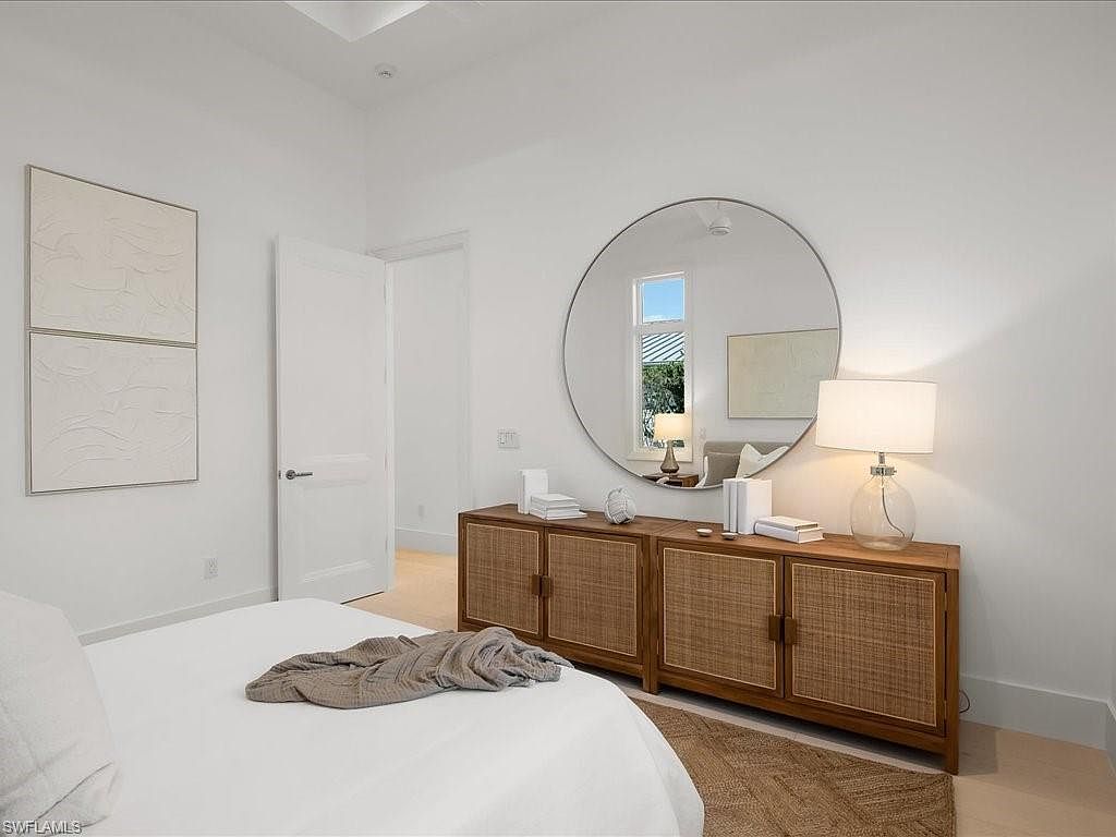 This is a bright and airy bedroom featuring a large round mirror above a wooden cabinet with woven door panels. The room is painted in a clean white, enhancing the natural light. A bed with white linens and a gray throw is partially visible in the foreground, suggesting a comfortable and inviting space.