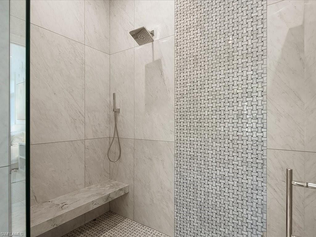 This is a well-appointed primary bathroom featuring a walk-in shower with marble-like tile walls and a decorative mosaic tile accent. The shower includes a rainfall showerhead, a handheld sprayer, and a built-in bench. The floor is covered in a small, patterned tile, and a glass enclosure partially surrounds the shower area, creating a luxurious and modern spa-like atmosphere.