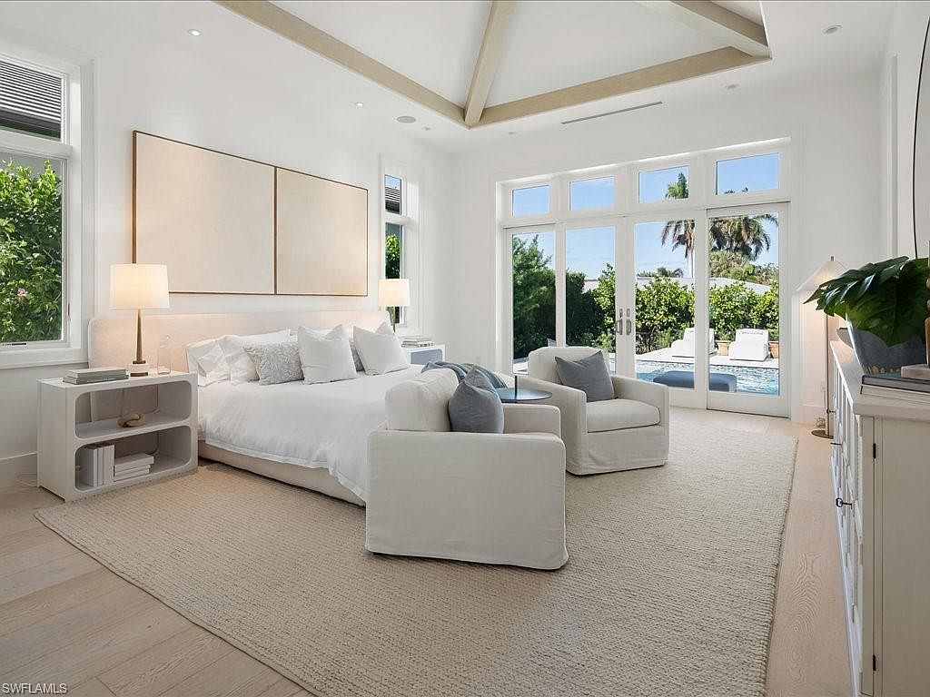 This is a bright and airy primary bedroom featuring a neutral color palette. The room includes a large bed with white linens, two armchairs, and a side table with a lamp. Large glass doors provide access to an outdoor area, likely a pool or patio, enhancing the indoor-outdoor flow.