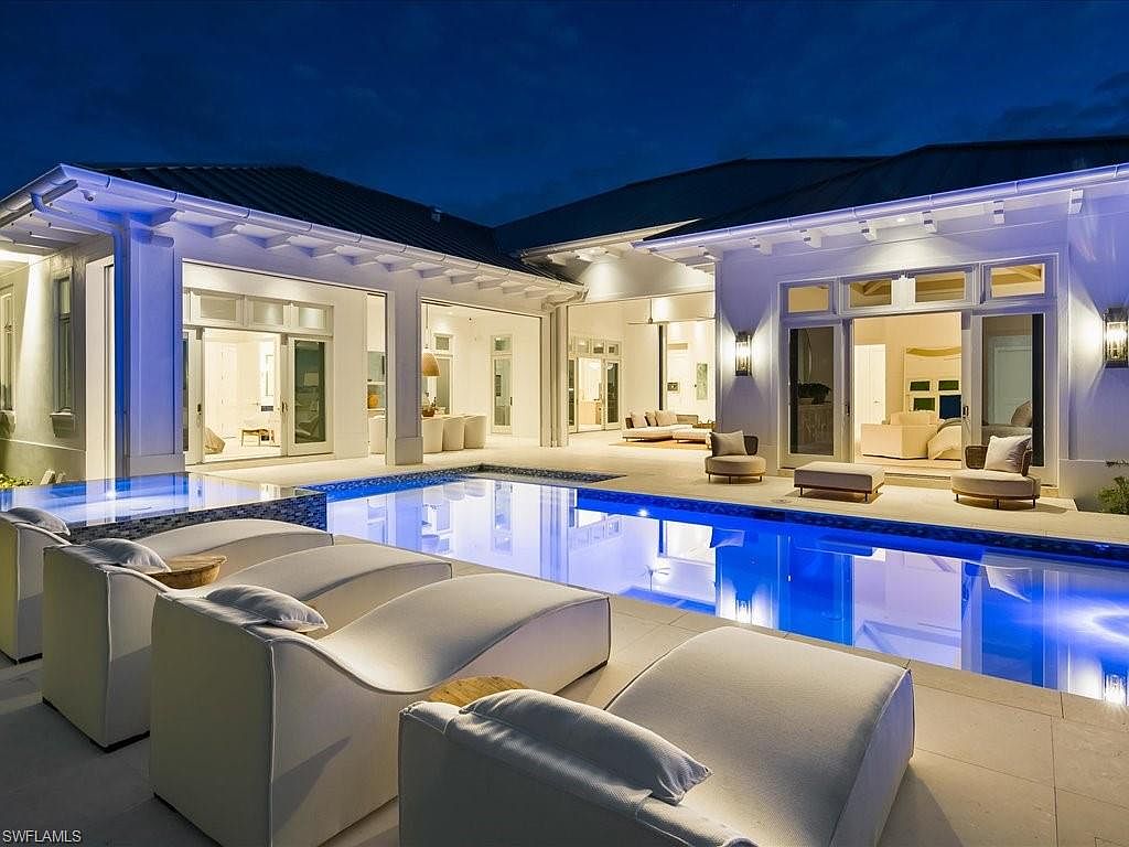 This image showcases a luxurious outdoor pool area at night, featuring a modern architectural style with white exterior walls and ample lighting. The pool is surrounded by comfortable lounge chairs, creating an inviting and relaxing atmosphere. Large glass doors open to the interior, seamlessly blending indoor and outdoor living spaces.