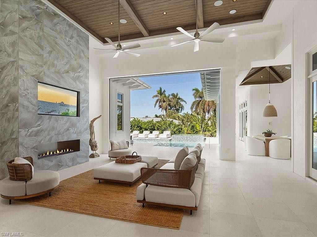 This is a bright and airy living room featuring a modern fireplace with a large stone surround and a flat-screen TV. The room is furnished with comfortable seating, including a sofa and armchairs, arranged around a jute rug. Large sliding glass doors open to an outdoor pool area, creating a seamless indoor-outdoor living experience.