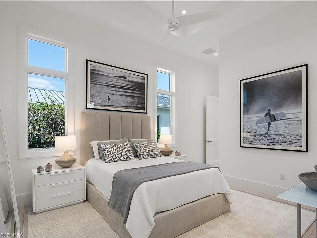 This is a bright and airy primary bedroom featuring a neutral color palette. The room is well-lit with natural light from the windows, complemented by recessed lighting and lamps on the nightstands. The bed has a padded headboard and is adorned with decorative pillows and a throw blanket, while artwork hangs on the walls, adding a touch of sophistication.
