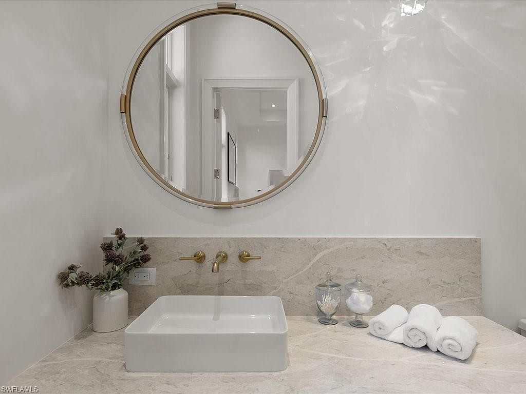 This is a well-lit bathroom featuring a modern, square-shaped white sink and gold-toned fixtures. A round mirror hangs above the sink, reflecting a glimpse into another room. The countertop and backsplash are made of a light-colored stone, and decorative elements like rolled towels, glass jars, and a small floral arrangement add a touch of elegance.