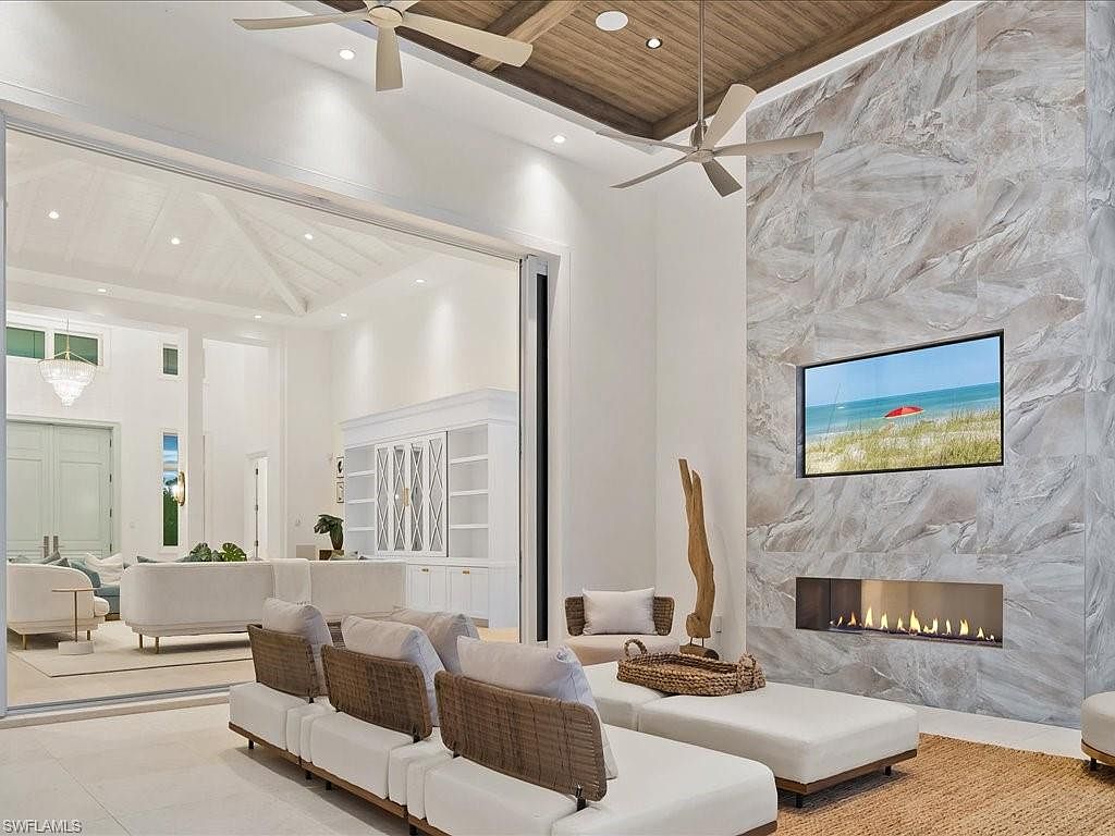 This is a bright and airy living room featuring a modern design with a neutral color palette. The room includes a large sectional sofa, a fireplace with a marble surround, and a flat-screen television. Large sliding glass doors open to an outdoor area, creating a seamless indoor-outdoor living experience.