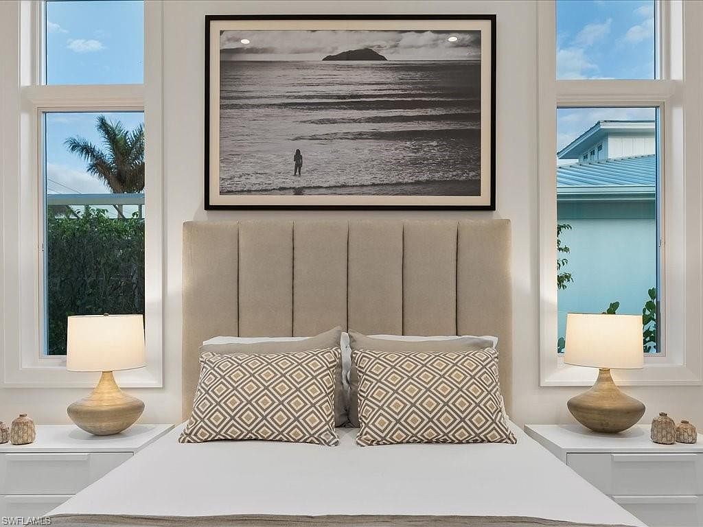 This is a well-lit primary bedroom featuring a neutral color palette. The focal point is a large upholstered headboard with a black and white framed picture above it. Two matching bedside tables with lamps flank the bed, which is adorned with decorative pillows and a white comforter, creating a serene and inviting atmosphere.