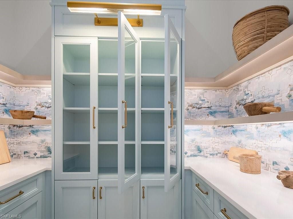 This interior shot showcases a well-organized pantry with light blue cabinetry and gold hardware. Glass-front cabinets display shelving, while lower cabinets provide additional storage. The pantry features decorative wallpaper and countertop space, creating a stylish and functional area.
