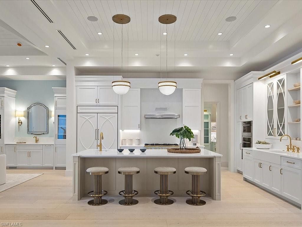 This is a bright and spacious kitchen featuring white cabinetry, a large center island with seating, and modern pendant lighting. The kitchen is well-equipped with stainless steel appliances and features a clean, contemporary design. The open layout and neutral color palette create an inviting and luxurious atmosphere.