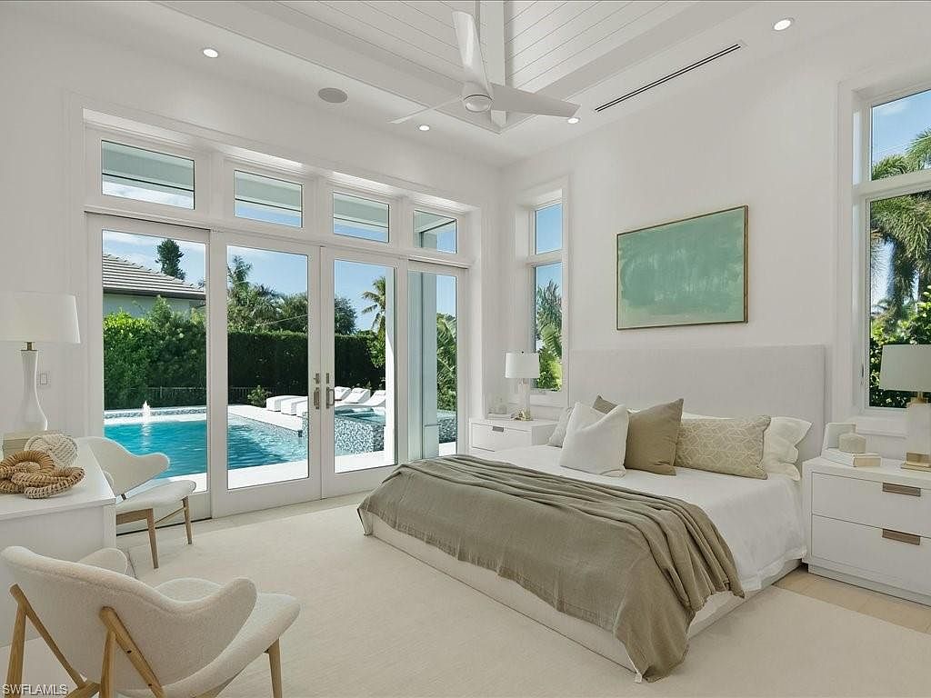 This is a bright and airy primary bedroom with a modern aesthetic. The room features a large bed with neutral-toned bedding, flanked by white nightstands and lamps. Large glass doors offer views of the pool and outdoor area, creating a seamless indoor-outdoor living experience.
