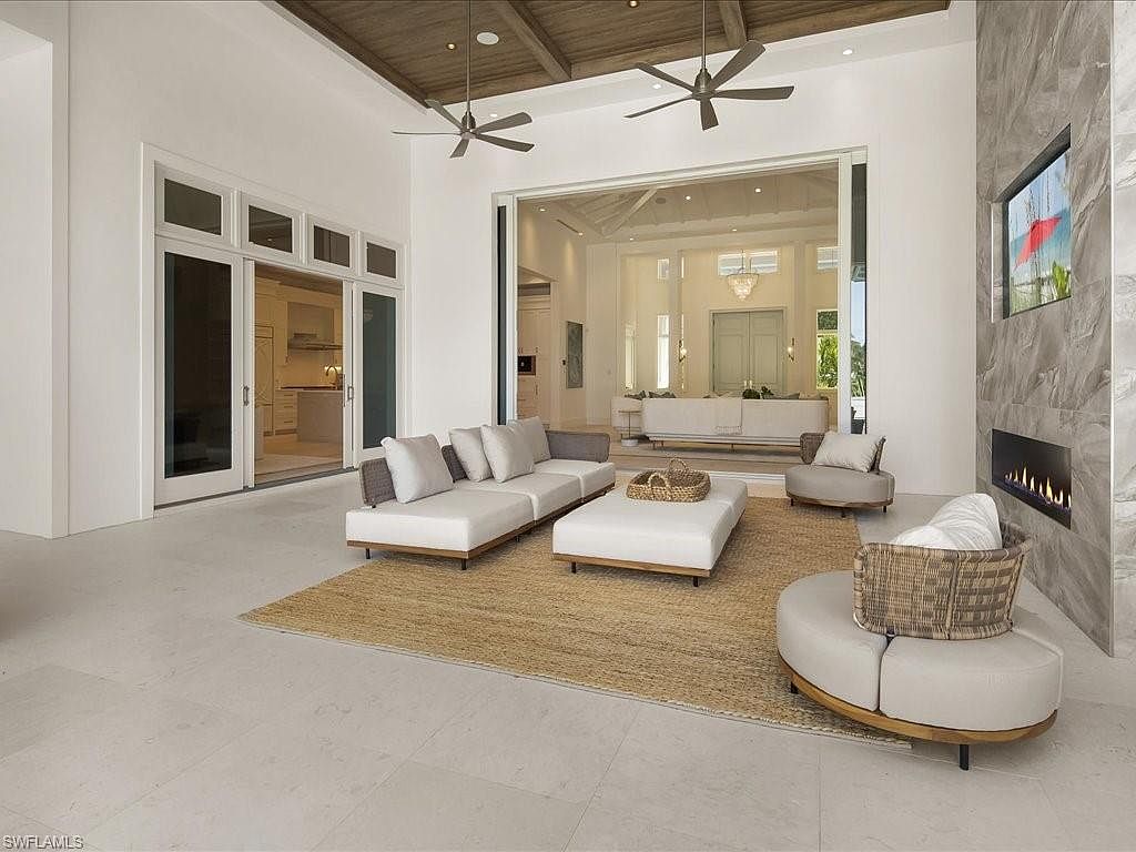 This is a bright and airy living room featuring a neutral color palette with white walls and light tile flooring. The room is furnished with modern white sofas, a large ottoman, and a round chair, all arranged on a jute rug. A fireplace with a stone surround and a large sliding glass door leading to another living space enhance the room's appeal, while ceiling fans provide air circulation.