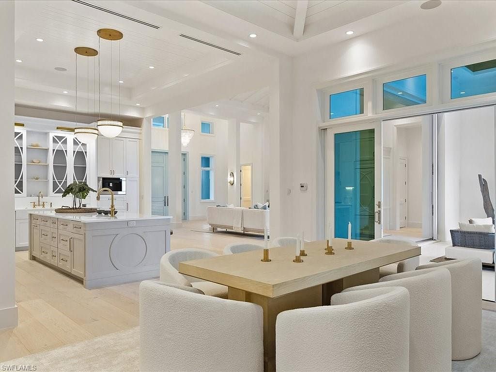 This interior shot showcases a bright and airy dining area adjacent to the kitchen and living spaces. The dining table, surrounded by plush white chairs, is the focal point, complemented by modern pendant lighting above the kitchen island. Large windows and sliding glass doors provide ample natural light, enhancing the open-concept design and creating a seamless flow between indoor and outdoor living.