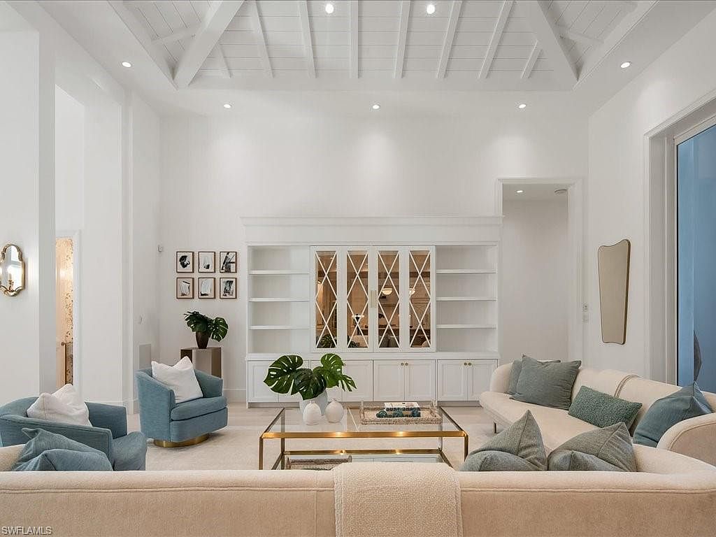 This is a bright and airy living room featuring high ceilings with white beams, creating a spacious and luxurious feel. The room is furnished with comfortable sofas and armchairs in neutral and blue tones, complemented by a glass-topped coffee table and a built-in shelving unit with mirrored accents. The overall aesthetic is modern and elegant, perfect for relaxation and entertaining.