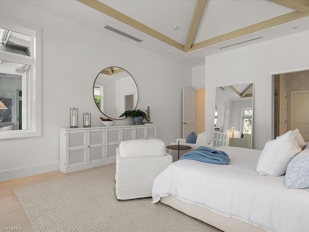 This is a bright and airy primary bedroom featuring a large bed with white linens and blue accents. The room includes a white dresser with a round mirror above it, decorative candles, and a plant. The ceiling has exposed wooden beams, and there is a doorway leading to another room, creating a sense of spaciousness and luxury.