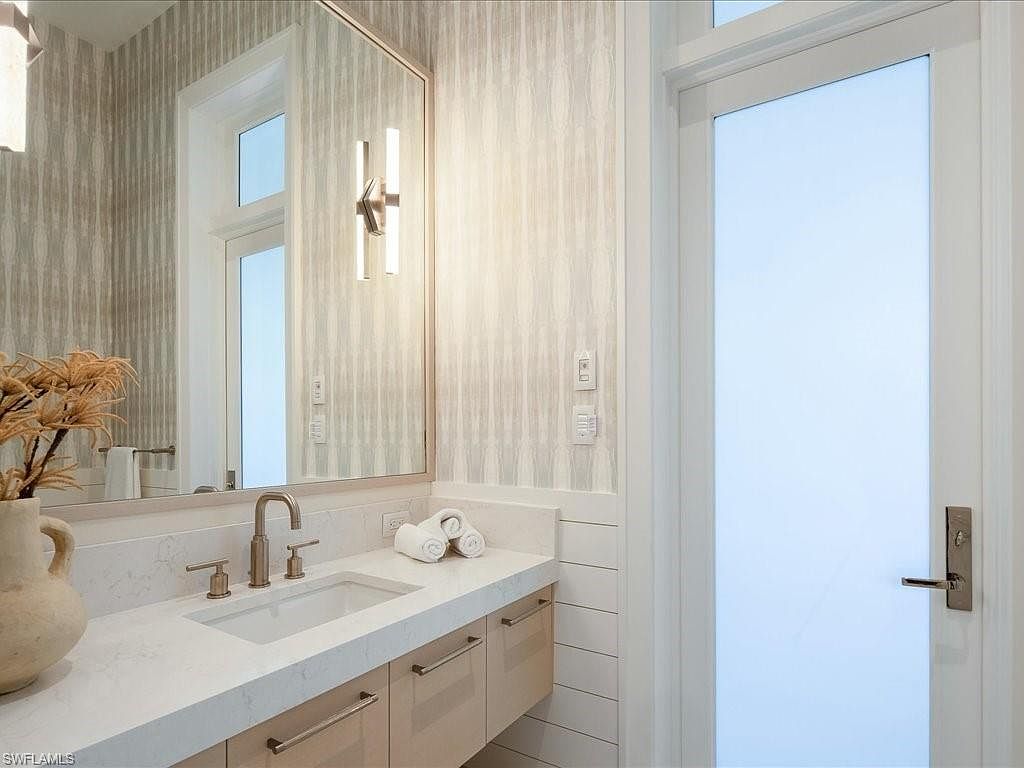 This is a well-lit bathroom featuring a modern vanity with light wood cabinetry and a white countertop. A large mirror reflects the space, and the walls are adorned with a subtle patterned wallpaper. The room has a clean and contemporary aesthetic, enhanced by the white door and trim.