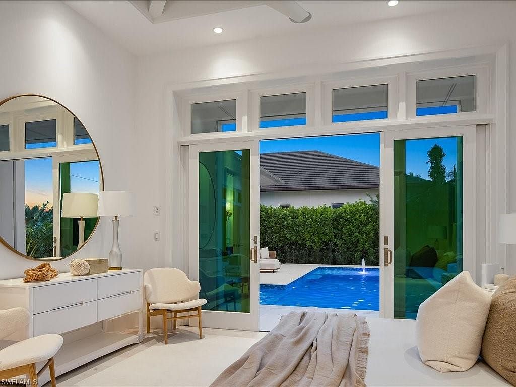 This is a bright and airy primary bedroom with a view of a private pool. The room features white walls, a large round mirror, and sliding glass doors that open to the outdoor pool area. The decor is minimalist and modern, creating a serene and luxurious atmosphere.