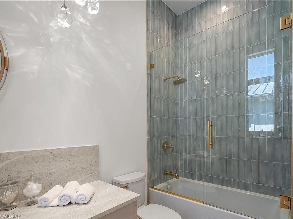This is a well-lit bathroom featuring a modern design with a glass-enclosed shower and a white toilet. The shower has vertically stacked blue-grey tiles and gold fixtures, including a rainfall showerhead. The vanity area includes a light-colored countertop with rolled white towels and decorative jars, creating a clean and inviting space.