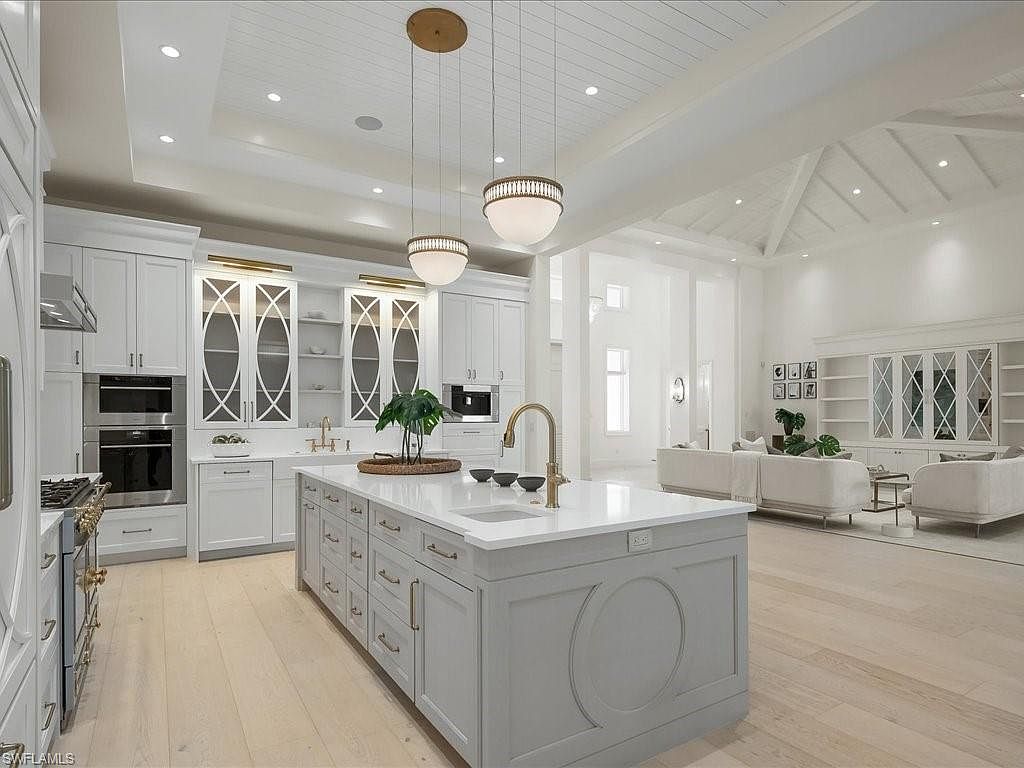 This is a bright and spacious kitchen that seamlessly transitions into a living area. The kitchen features a large island with a white countertop, gray cabinetry, and gold hardware, complemented by pendant lighting. The living area includes comfortable seating and built-in shelving, creating an open and inviting atmosphere with a modern aesthetic.