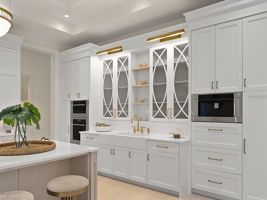 This is a bright and modern kitchen featuring white cabinetry with gold hardware, glass-front cabinets with decorative mullions, and built-in stainless steel appliances. The kitchen island has a white countertop and seating, complemented by a light wood floor. The overall impression is clean, elegant, and well-designed, perfect for a high-end real estate listing.