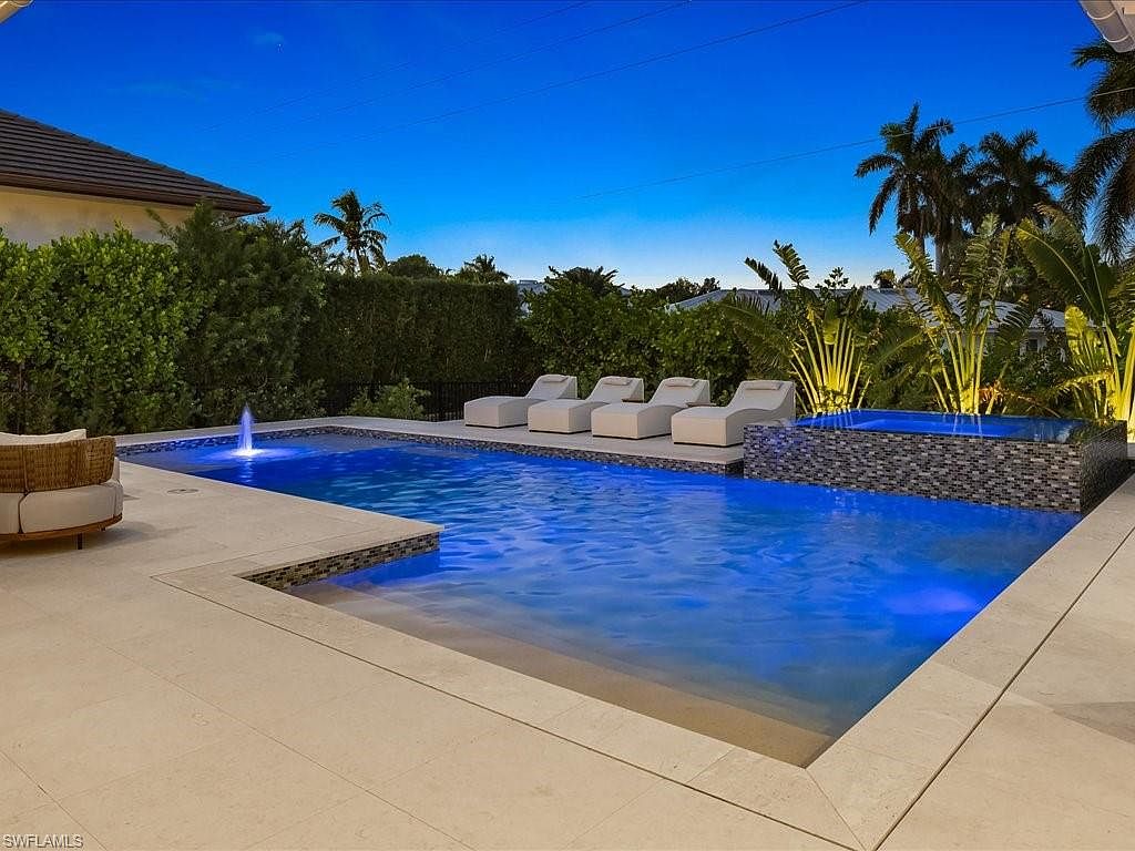 This image showcases a luxurious outdoor pool and spa area at dusk. The pool features a modern design with a shallow entry area and a fountain, while the spa is elevated and tiled with mosaic patterns. Lounge chairs are arranged poolside, and tropical landscaping adds to the serene and upscale ambiance, making it an inviting space for relaxation and entertainment.