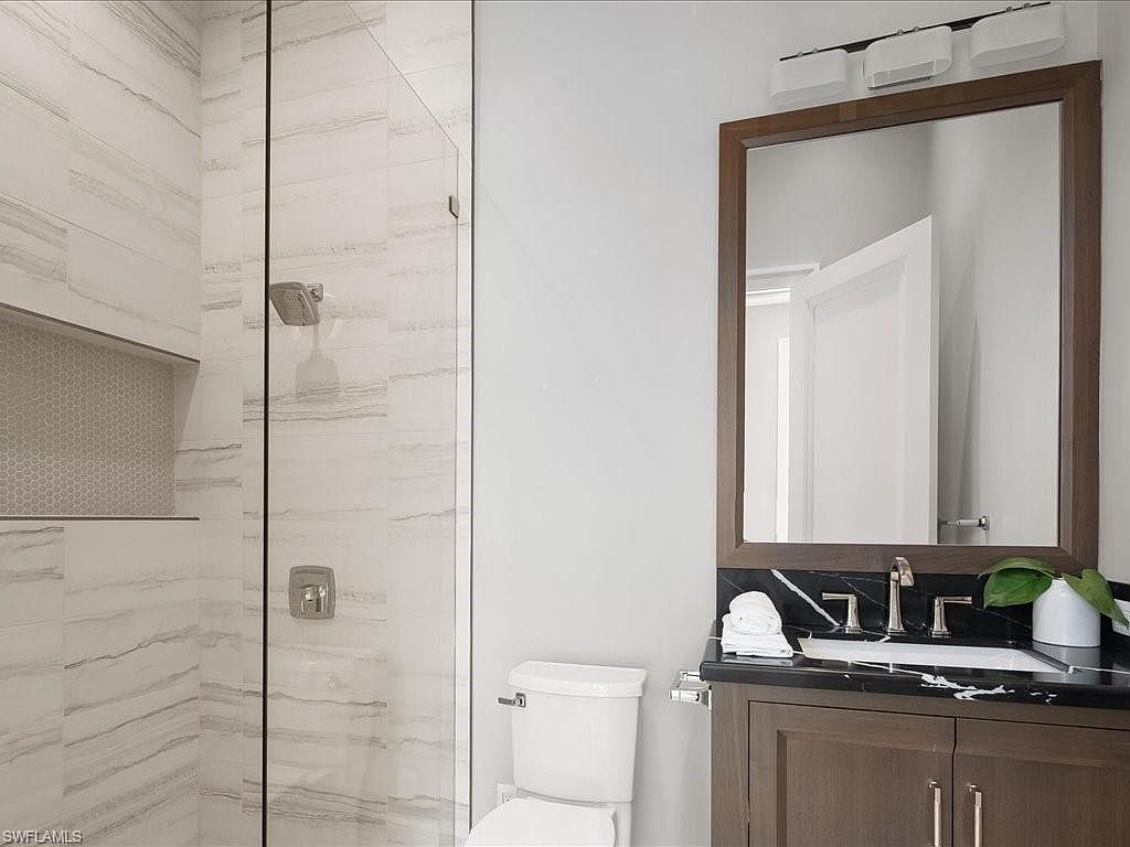 This is a well-lit bathroom featuring a shower with glass doors and tiled walls, a toilet, and a vanity with a dark wood cabinet and a black countertop. A framed mirror hangs above the vanity, reflecting a glimpse of a white door. The overall impression is clean and modern.