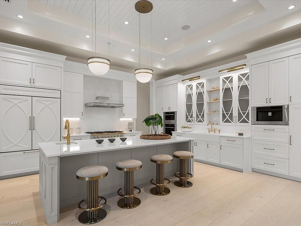 This is a bright and spacious kitchen featuring white cabinetry, stainless steel appliances, and a large center island with seating. The kitchen is illuminated by pendant lighting and recessed lights, creating a warm and inviting atmosphere. The perspective is a wide shot, capturing the entire kitchen layout and highlighting its modern design.