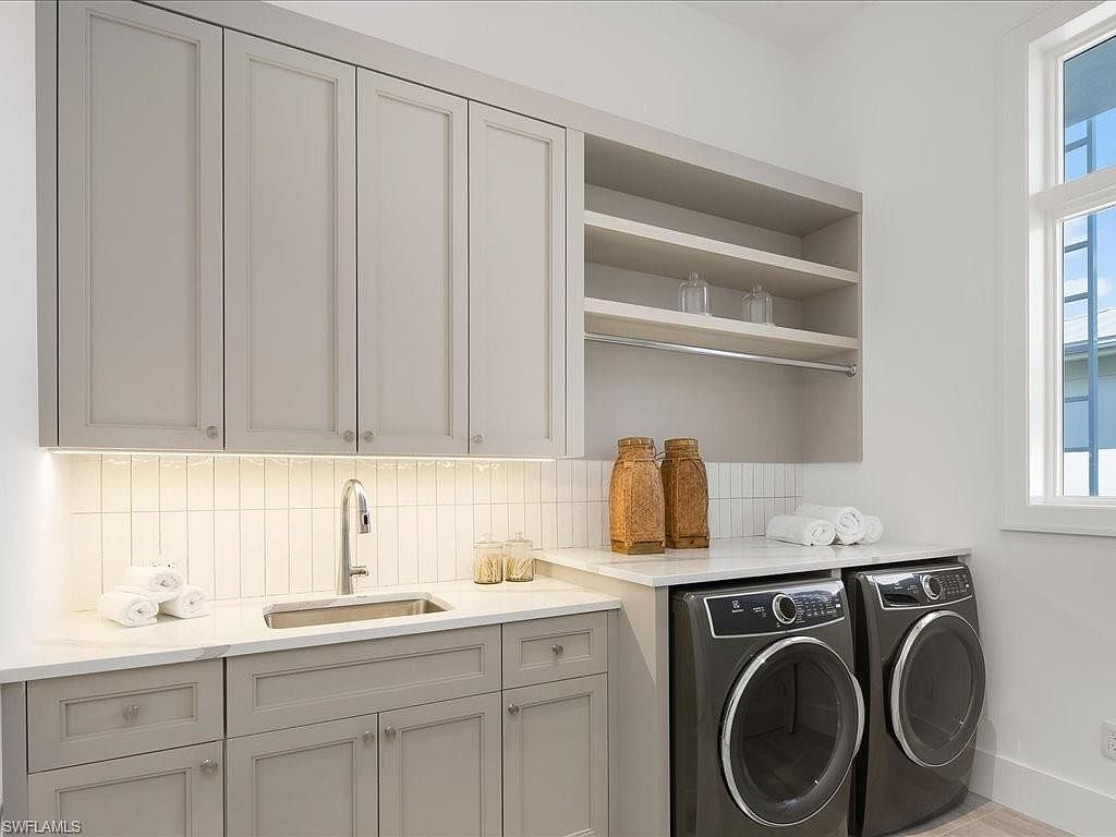 This is a well-organized laundry room featuring gray cabinetry with silver hardware, a white countertop, and a stainless steel sink. A subway tile backsplash adds a touch of modern style, while open shelves provide additional storage. The room also includes a front-loading washer and dryer set, creating a functional and aesthetically pleasing space.