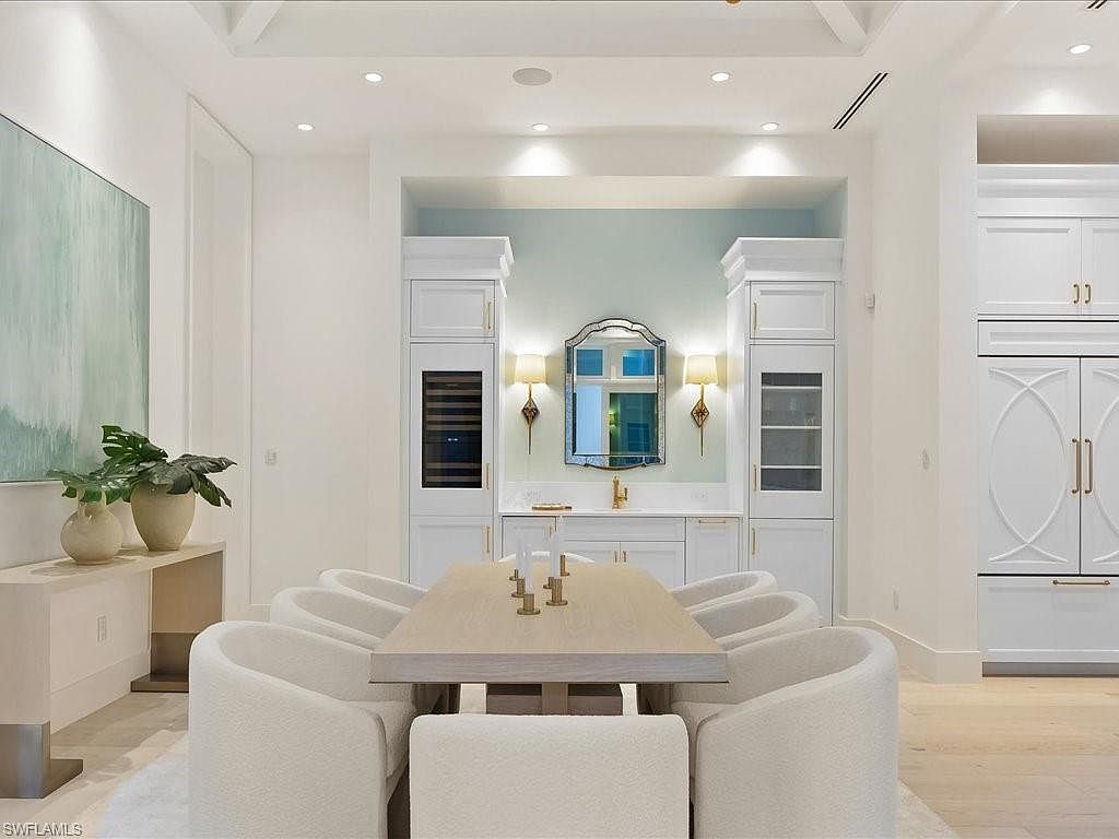 This is an interior shot of a dining room featuring a light wood table surrounded by white upholstered chairs. The room has white walls and cabinetry, with a built-in bar area in the background featuring a mirror and sconces. The overall aesthetic is bright, clean, and modern, creating an inviting space for dining and entertaining.