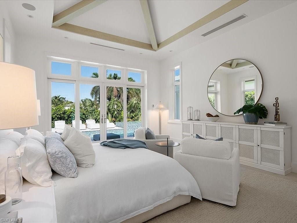 This is a bright and airy primary bedroom featuring a large bed with white linens and decorative pillows. The room has a modern coastal style with white walls, light-colored flooring, and a large window offering a view of the pool and lush greenery. A round mirror and a white cabinet add to the room's elegant and serene atmosphere.
