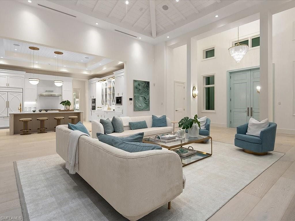 This is an interior shot of a luxurious living room that seamlessly blends into a modern kitchen. The living room features plush sofas with blue accent pillows, a glass coffee table, and a light-colored rug, while the kitchen boasts white cabinetry, a large island with bar stools, and pendant lighting. The overall impression is one of spaciousness, elegance, and contemporary design, enhanced by the high ceilings and natural light.