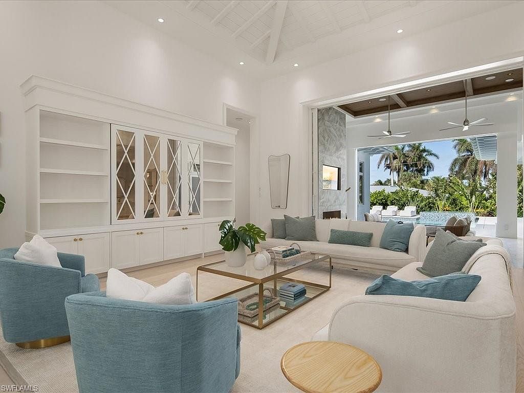 This is a bright and airy living room featuring a large white sectional sofa with blue accent pillows, two blue armchairs, and a glass coffee table. A built-in white cabinet with glass doors provides ample storage and display space. The room opens to an outdoor patio and pool area, creating a seamless indoor-outdoor living experience.