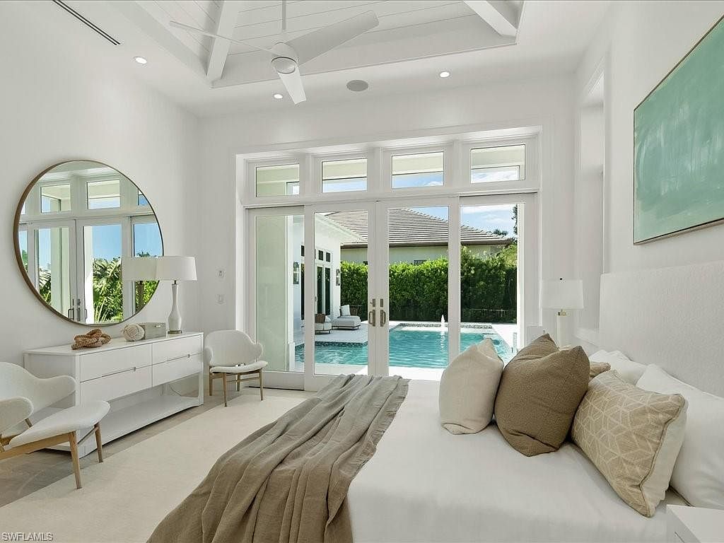 This is a bright and airy primary bedroom featuring a large bed with neutral-toned pillows and a throw blanket. A round mirror hangs above a white dresser, and sliding glass doors offer a view of the pool area. The room is decorated in a minimalist style with white walls and light-colored furniture, creating a serene and luxurious atmosphere.