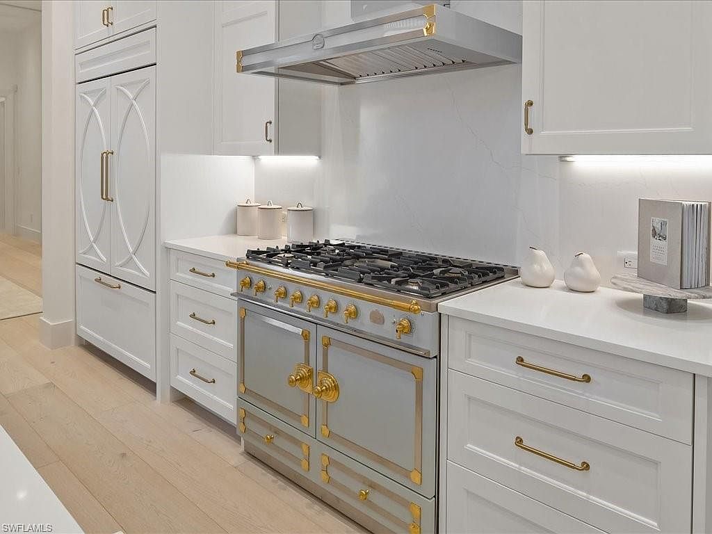 This is a bright and elegant kitchen featuring white cabinetry with gold hardware, a professional-grade range with a custom hood, and light hardwood flooring. The countertops are a clean white, complementing the overall sophisticated design. The perspective is a medium shot, showcasing the range and surrounding cabinetry.