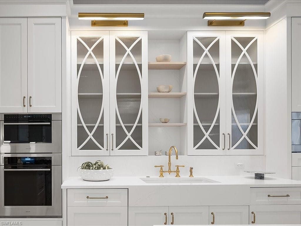 This is a well-lit kitchen featuring white cabinetry with glass-fronted upper cabinets and gold hardware. The countertops are white quartz, and a farmhouse sink is centered beneath the cabinets. Stainless steel appliances are built into the cabinetry, creating a seamless and modern look.