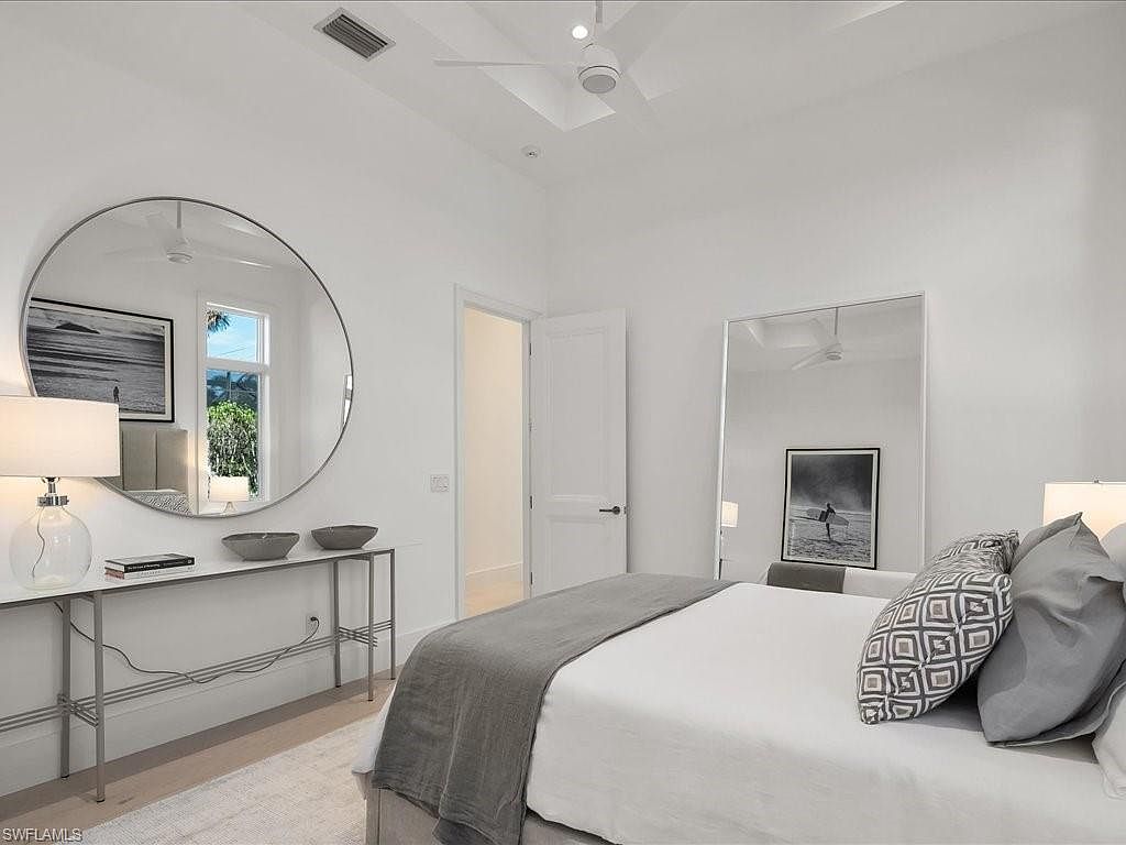 This is a bright and airy primary bedroom featuring a neutral color palette. The room includes a large round mirror above a console table, a bed with decorative pillows and a throw blanket, and a full-length mirror reflecting the room's details. The overall impression is clean, modern, and serene, creating a relaxing atmosphere.