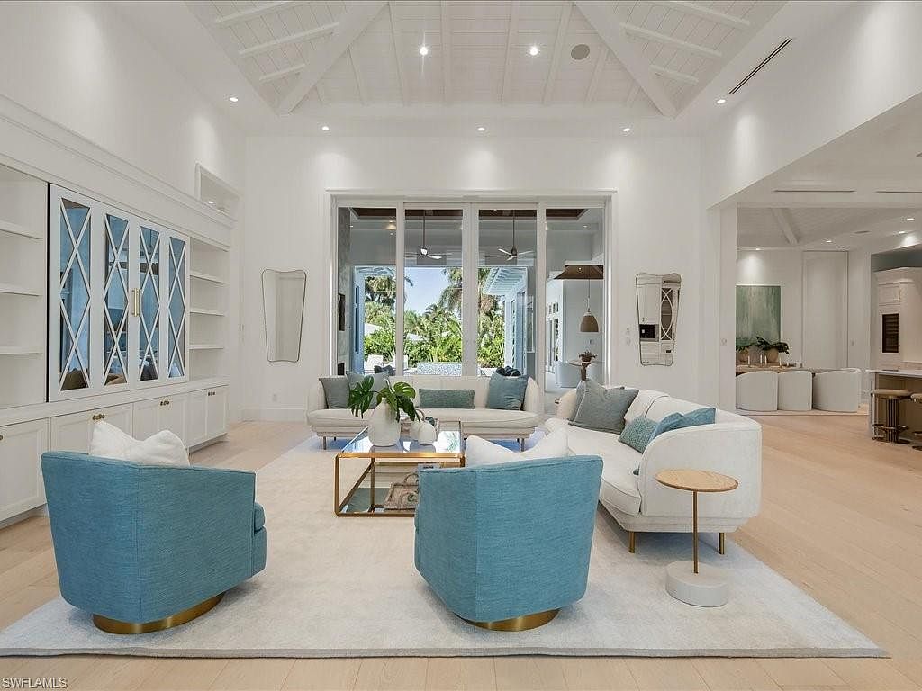 This is a bright and airy living room featuring a neutral color palette with pops of blue. The room is furnished with two white sofas, two blue armchairs, and a glass coffee table with gold accents. Large windows provide ample natural light and a view of the outdoors, enhancing the spacious feel of the room.