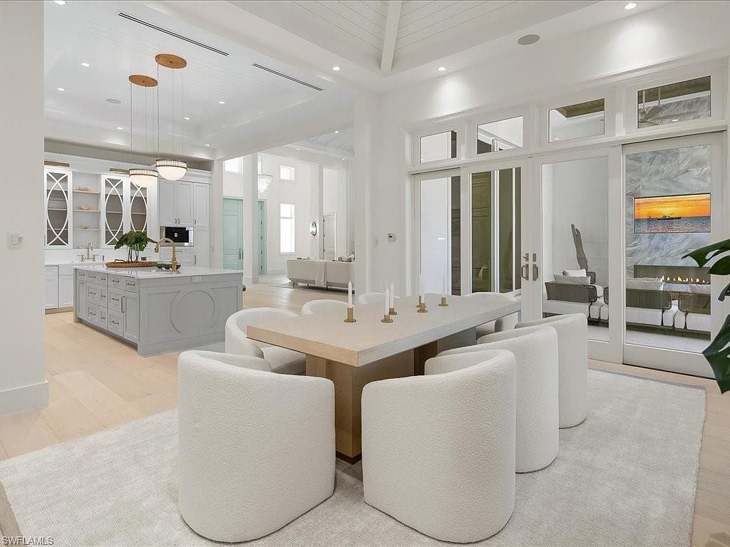 This interior shot showcases a bright and airy dining room with a modern aesthetic. The room features a large, light-colored dining table surrounded by plush, white chairs, set upon a light-colored rug. Sliding glass doors provide ample natural light and a view into another room, while a glimpse of the kitchen can be seen in the background, creating an open and inviting atmosphere.