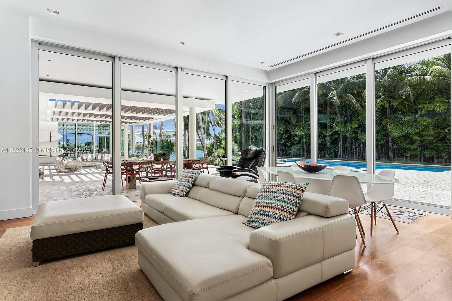 This bright and airy living room features a large, light-colored sectional sofa and a matching ottoman, centered on a neutral area rug. The space is defined by expansive floor-to-ceiling glass walls that provide seamless indoor-outdoor flow to a covered patio and a lush, tropical backyard with a swimming pool. The interior design is minimalist and contemporary, with warm wood flooring and a dining area visible in the background, creating an inviting and open-concept atmosphere.