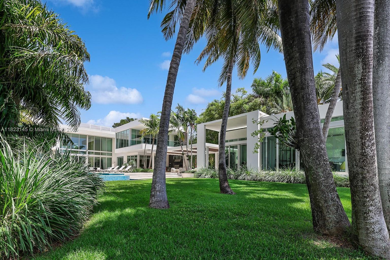 This modern, multi-level residence features a striking white facade with extensive floor-to-ceiling glass walls that seamlessly connect the interior living spaces to the outdoors. The property is set against a lush, manicured lawn framed by tall palm trees, with a sparkling swimming pool visible in the foreground. The architectural design emphasizes clean lines and an open, airy aesthetic, creating a sophisticated and tranquil retreat.