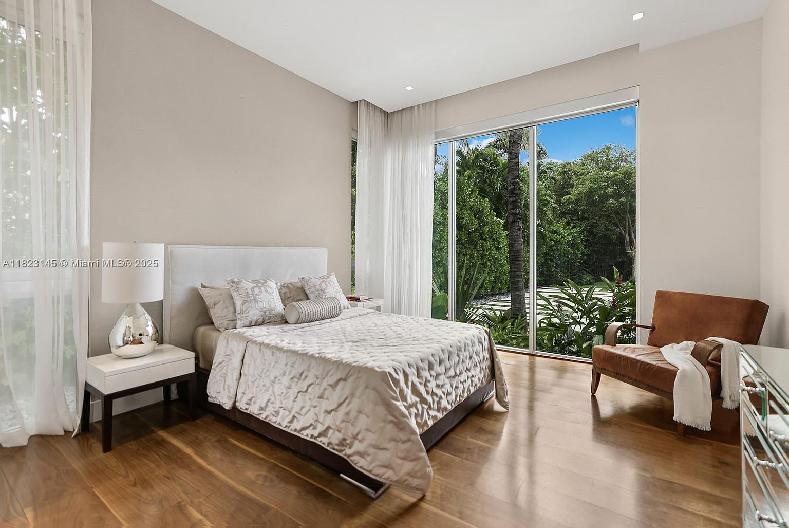 This bright and airy bedroom features warm wood flooring and a neutral color palette, centered around a plush bed with a tufted headboard. Large windows provide an expansive view of the lush, green outdoor landscape, filling the room with natural light. A comfortable leather armchair sits in the corner, creating a cozy reading nook that complements the serene and sophisticated atmosphere of the space.