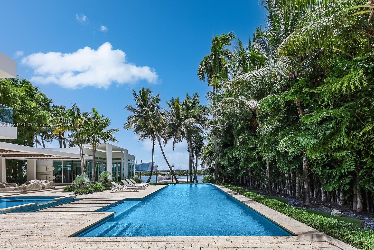 This luxurious outdoor space features a long, rectangular swimming pool set against a backdrop of lush palm trees and dense greenery. A modern, light-colored stone patio surrounds the pool, complemented by a covered lounge area with comfortable outdoor seating and a view of the water beyond. The scene captures a serene, resort-like atmosphere perfect for relaxation and entertaining.