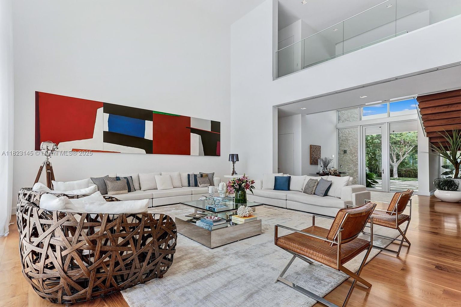 This expansive, double-height living room features a bright, open-concept design with white walls and warm wood flooring. The space is anchored by a large, abstract red and blue painting, a neutral sectional sofa, and a unique woven accent chair, all centered around a glass-topped coffee table. Large windows and glass doors provide a seamless connection to the outdoor greenery, while a glass-railed mezzanine overlooks the area from above.