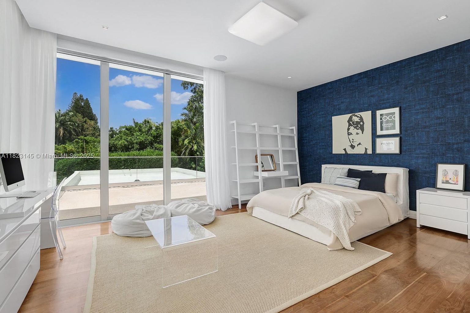 This bright and airy bedroom features a striking navy blue textured accent wall adorned with framed black-and-white art. A plush bed with neutral bedding sits on a large area rug, complemented by a white bookshelf and a minimalist desk setup. The room is bathed in natural light from expansive floor-to-ceiling windows that provide a seamless view of the outdoor terrace and lush greenery beyond.