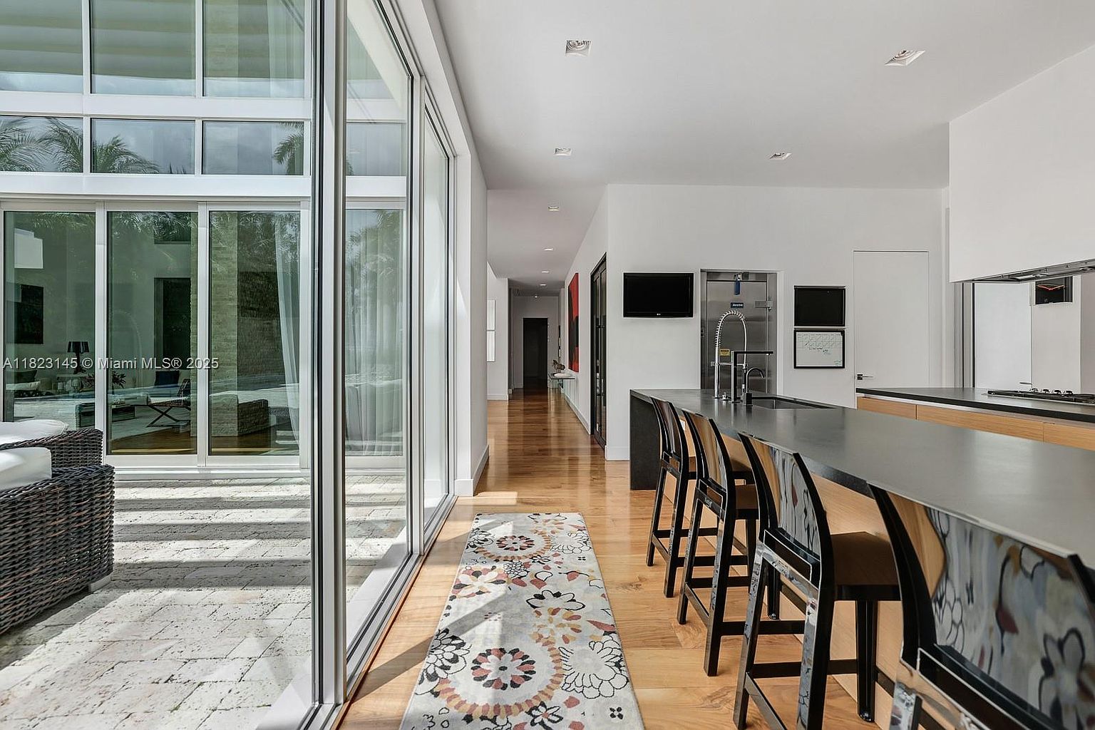 This modern kitchen features a long island with a dark countertop and bar seating, set against a backdrop of light wood cabinetry and a hallway leading to the rest of the home. Large, floor-to-ceiling glass sliding doors provide a seamless transition to an outdoor patio, filling the space with natural light. The interior design is sleek and minimalist, emphasizing clean lines and an open-concept layout.