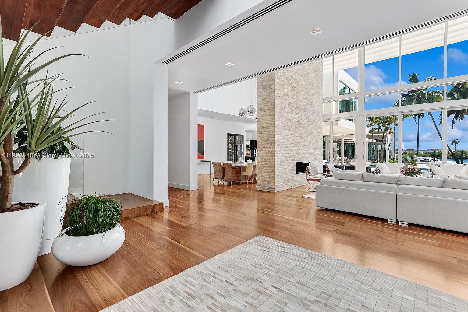 This expansive, open-concept living area features warm wood flooring and a bright, airy atmosphere created by large glass walls that open to an outdoor view. A prominent stone-clad chimney breast serves as a central architectural feature, while a neutral-toned sectional sofa provides comfortable seating. The perspective looks from a transition space with potted plants toward the main living area and a dining space in the background, emphasizing the home's spacious and modern design.