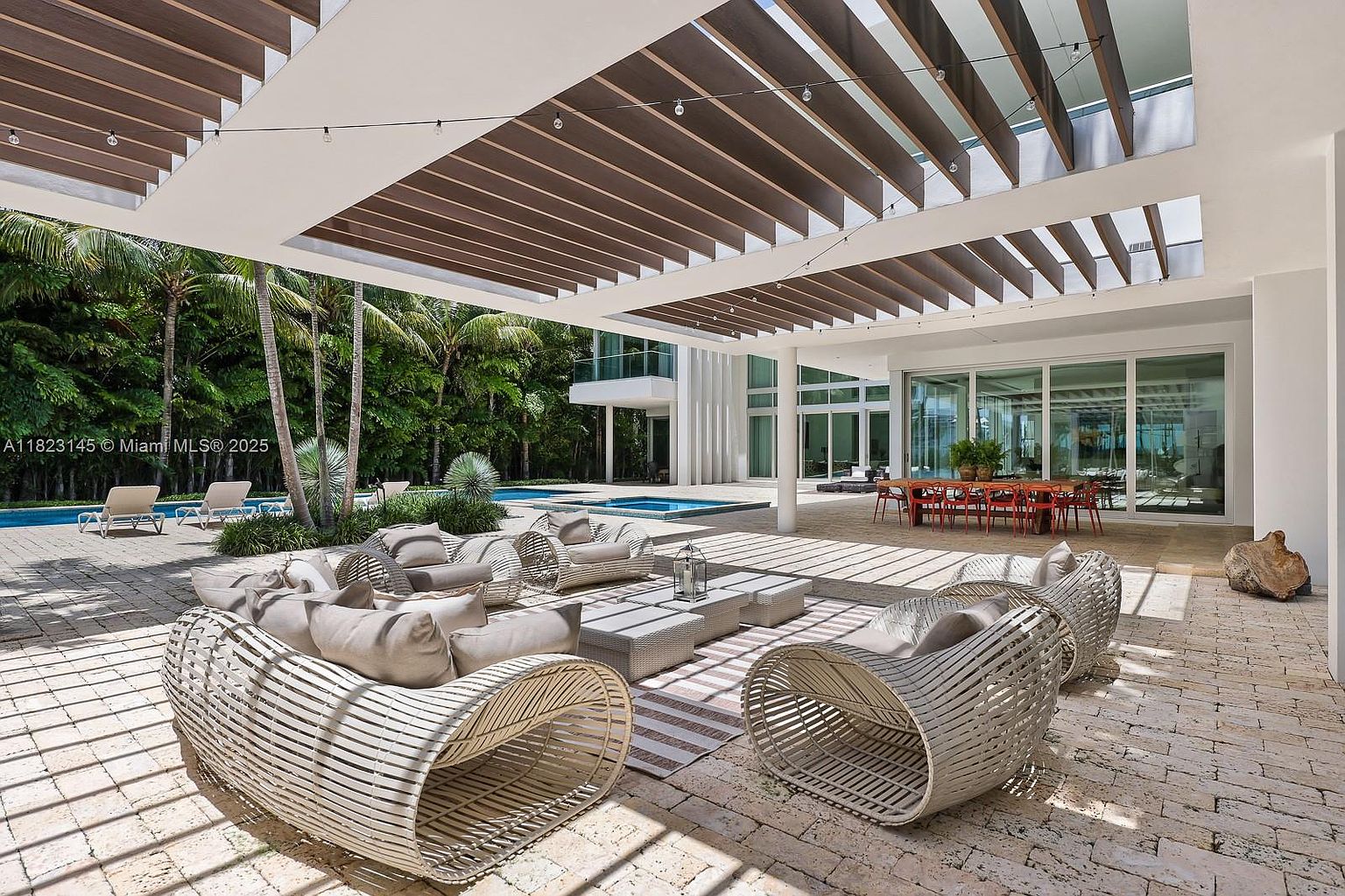 This expansive covered patio features a modern architectural design with a slatted wood-look ceiling and integrated lighting. The space is furnished with contemporary woven lounge seating and a dining area, all set against a backdrop of lush tropical landscaping and a swimming pool. The perspective captures the seamless transition between the indoor living area and the outdoor oasis, emphasizing a luxurious, resort-style atmosphere.