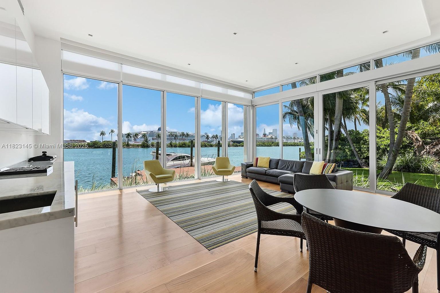 This bright, open-concept living area features expansive floor-to-ceiling windows that provide panoramic views of the waterfront and distant city skyline. The space is furnished with a sleek, dark leather sectional, two lime-green accent chairs, and a dining set, all anchored by a large, striped area rug on light wood flooring. The minimalist design emphasizes the connection between the indoor living space and the scenic outdoor environment.