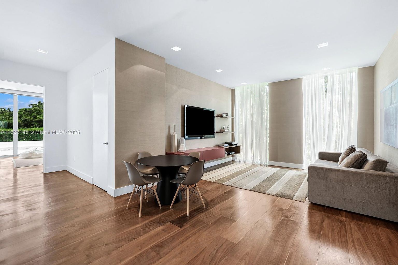 This modern living and dining area features warm wood flooring and a neutral color palette, creating a sophisticated and inviting atmosphere. A sleek, wall-mounted television sits above a long, low-profile console, while a contemporary dining set occupies the center of the room. Large windows draped in sheer curtains allow natural light to fill the space, and a doorway leads to an outdoor balcony area.